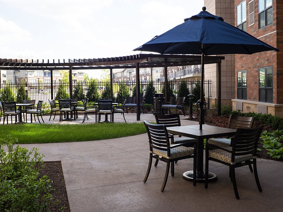 Apartment patio area with tables, chairs, a pergola, and a blue umbrella on a sunny day.