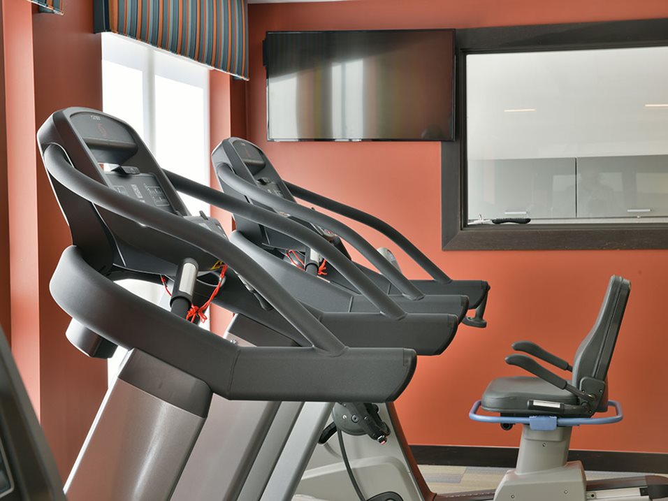 Three exercise machines in a brightly lit gym room with a wall-mounted television.