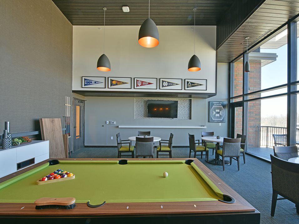 A billiards room with a green pool table, chairs, tables, and wall-mounted college banners.