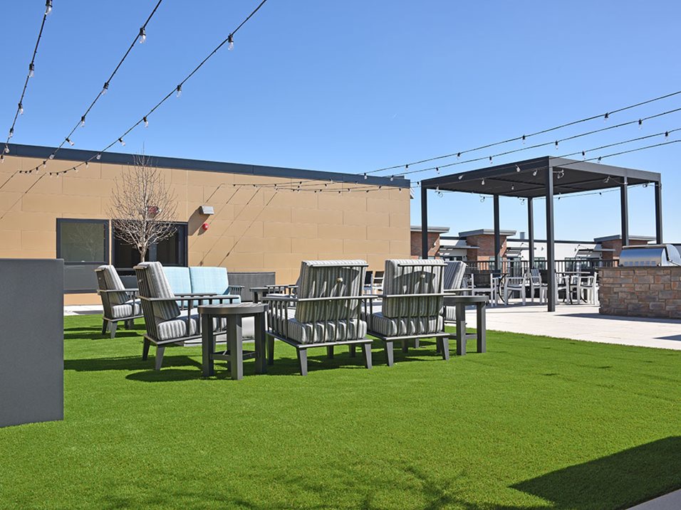 Rooftop lounge area with seating, green artificial grass, string lights, and a clear blue sky.