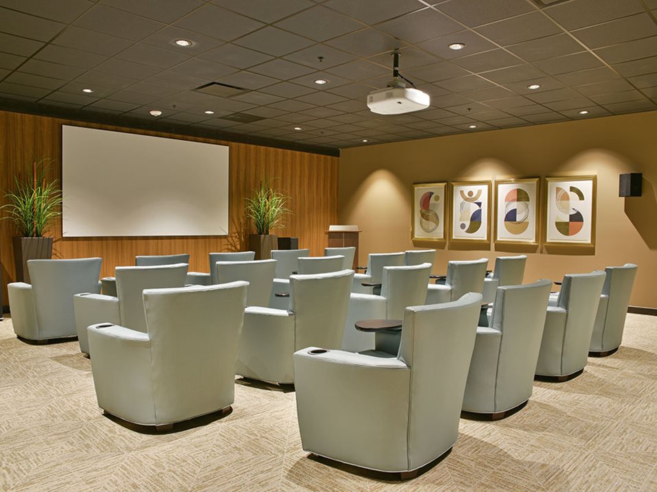 Small conference room with rows of light blue chairs facing a projector screen on a wooden wall.