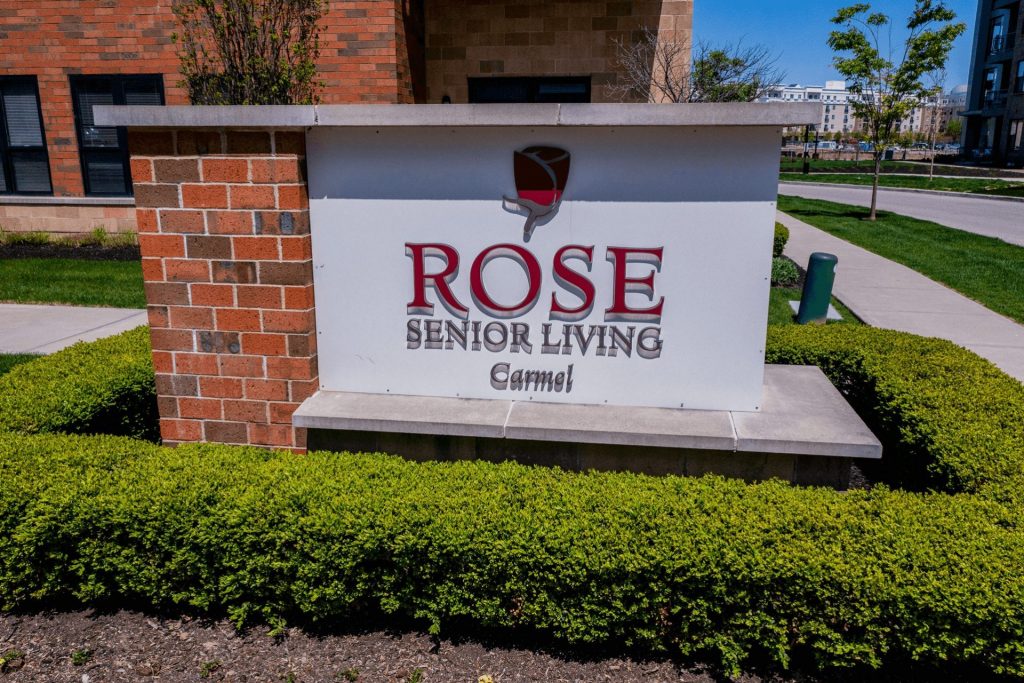 A sign for Rose Senior Living Carmel with greenery and a brick building in the background.