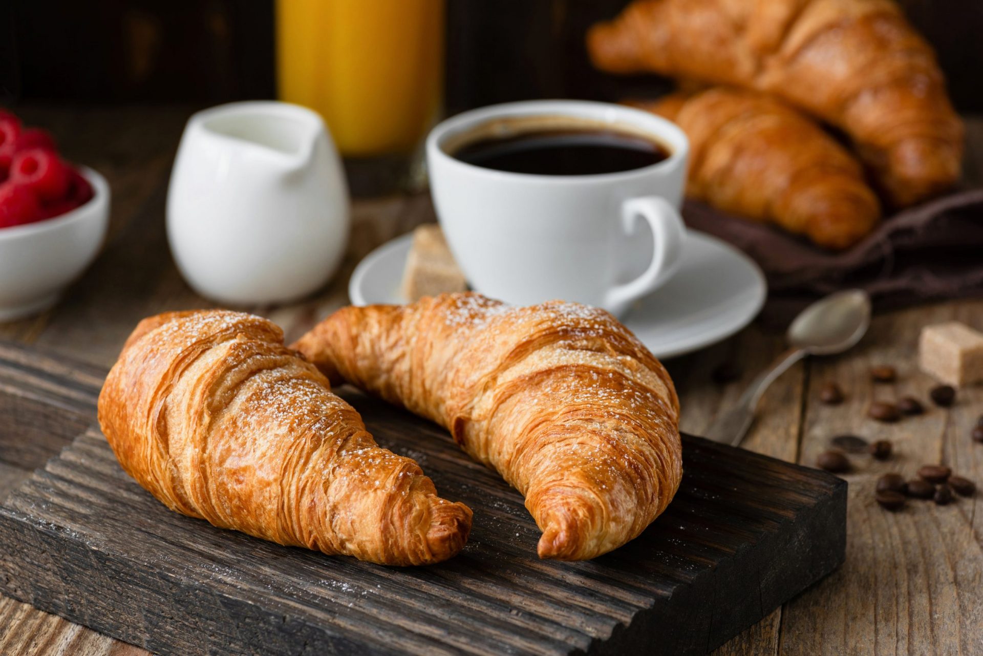 Two croissants on a wooden board alongside a cup of coffee, cream, and raspberries