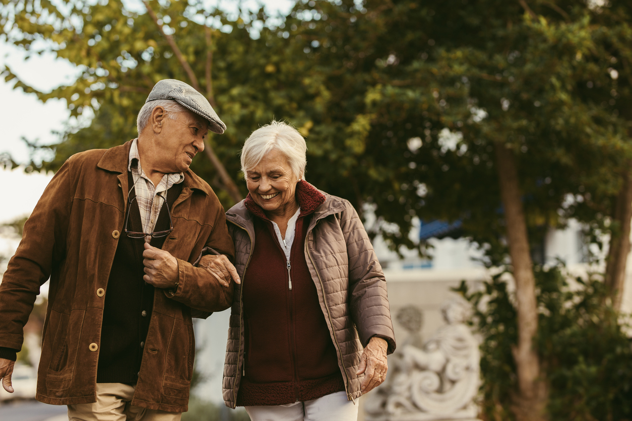 Elderly couple smiling and walking arm in arm in a park on a sunny day.