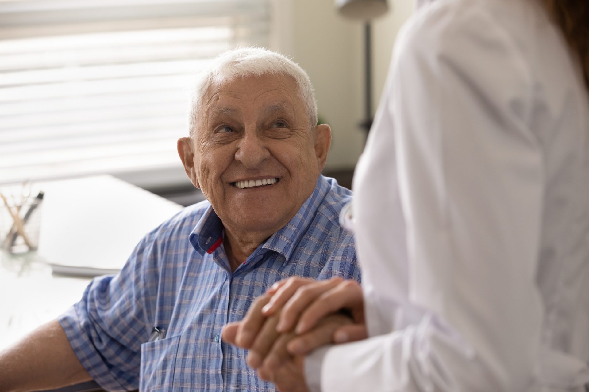 Elderly man smiling and holding hands with a doctor during a check-up in a brightly lit room.