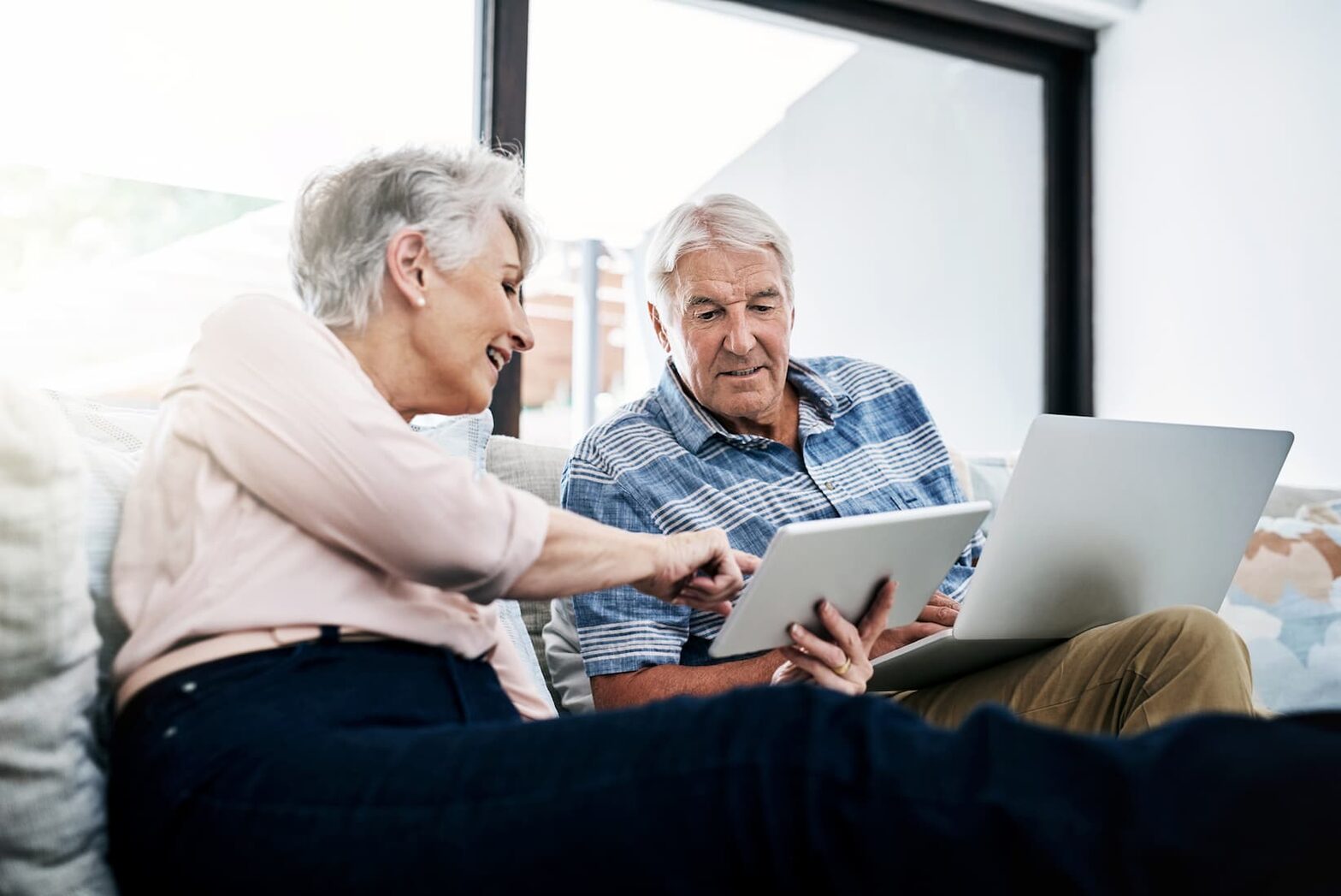 Senior man and woman sit on a couch and review information on a laptop and tablet screen together